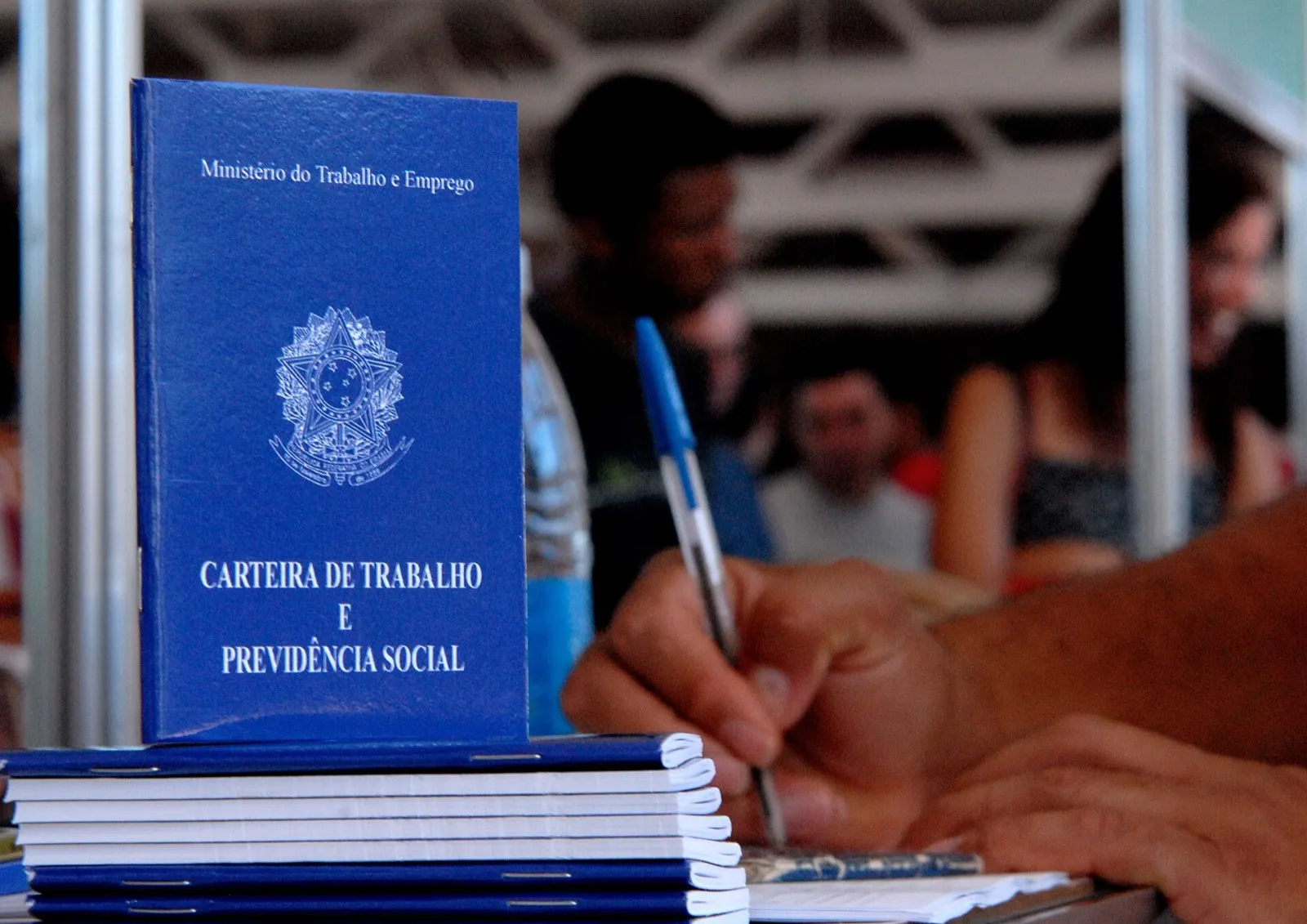Carteira de Trabalho e Previdência Social de capa azul, em pé sobre uma pilha de carteiras, com uma pessoa escrevendo ao fundo