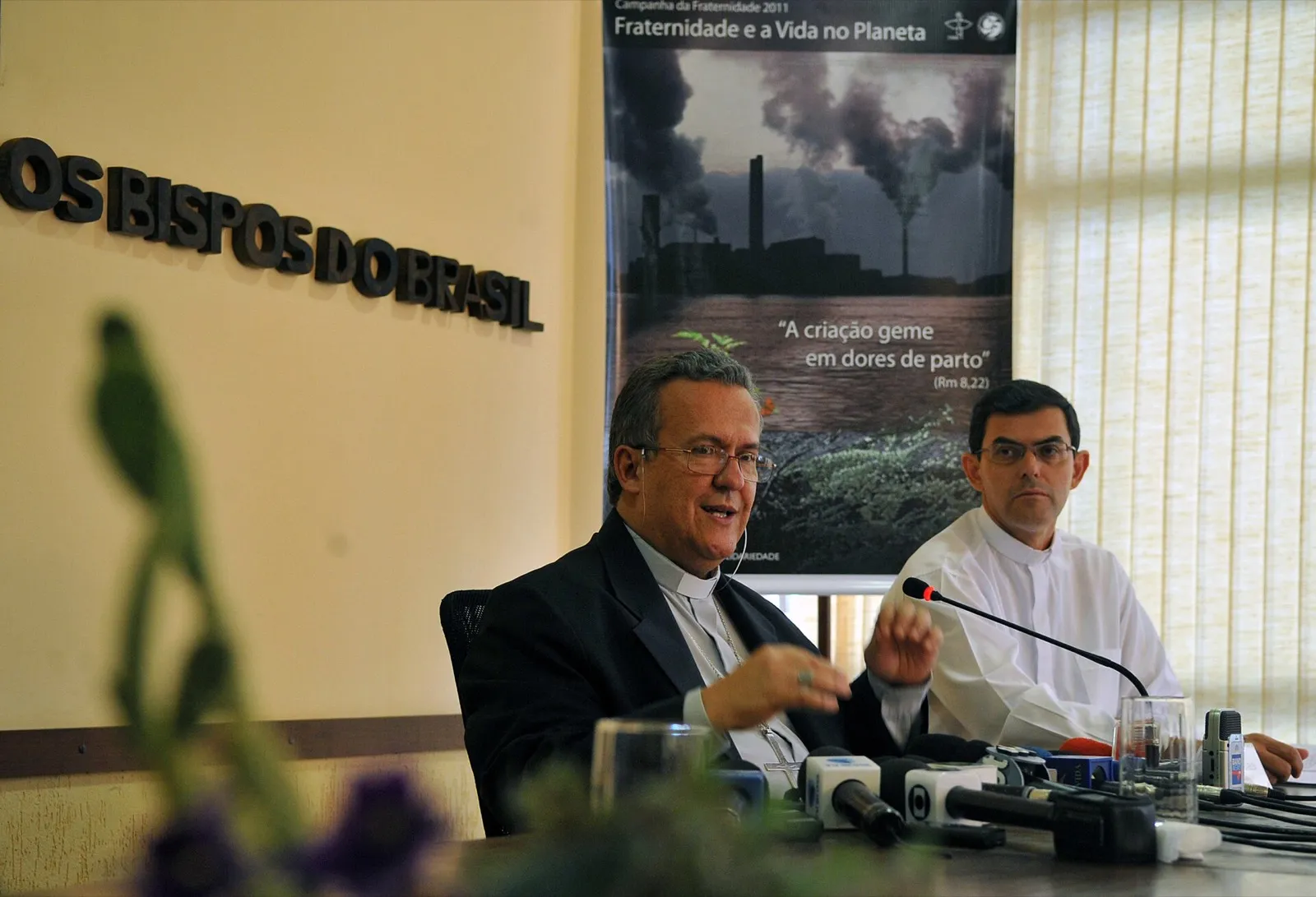 Dois religiosos sentados à mesa de uma coletiva, com microfones à frente e um cartaz da Campanha da Fraternidade 2011 ao fundo