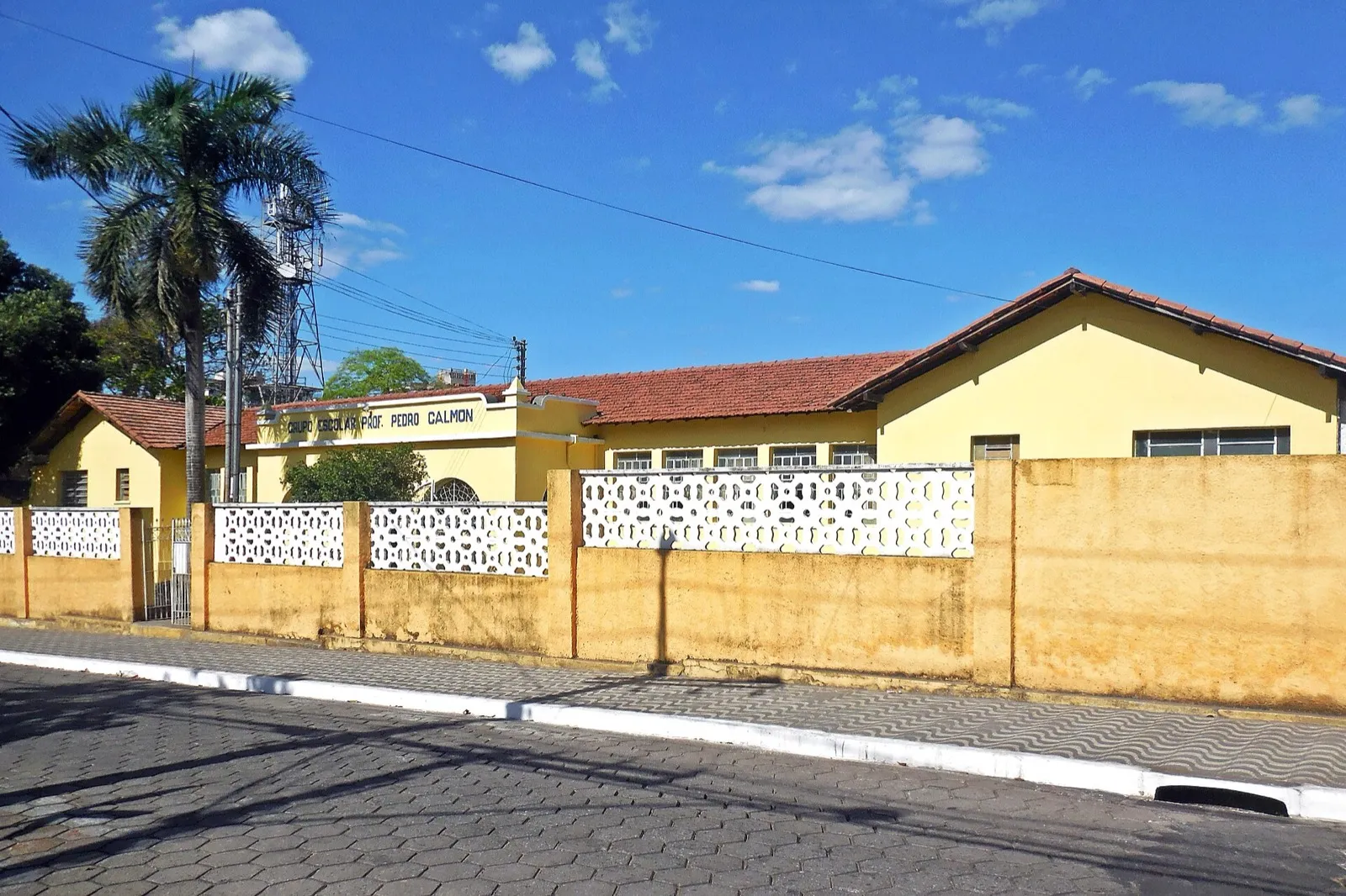 Prédio térreo amarelo de escola pública com telhado de barro, muro baixo com elementos vazados e a inscrição Grupo Escolar Prof. Pedro Calmon na fachada