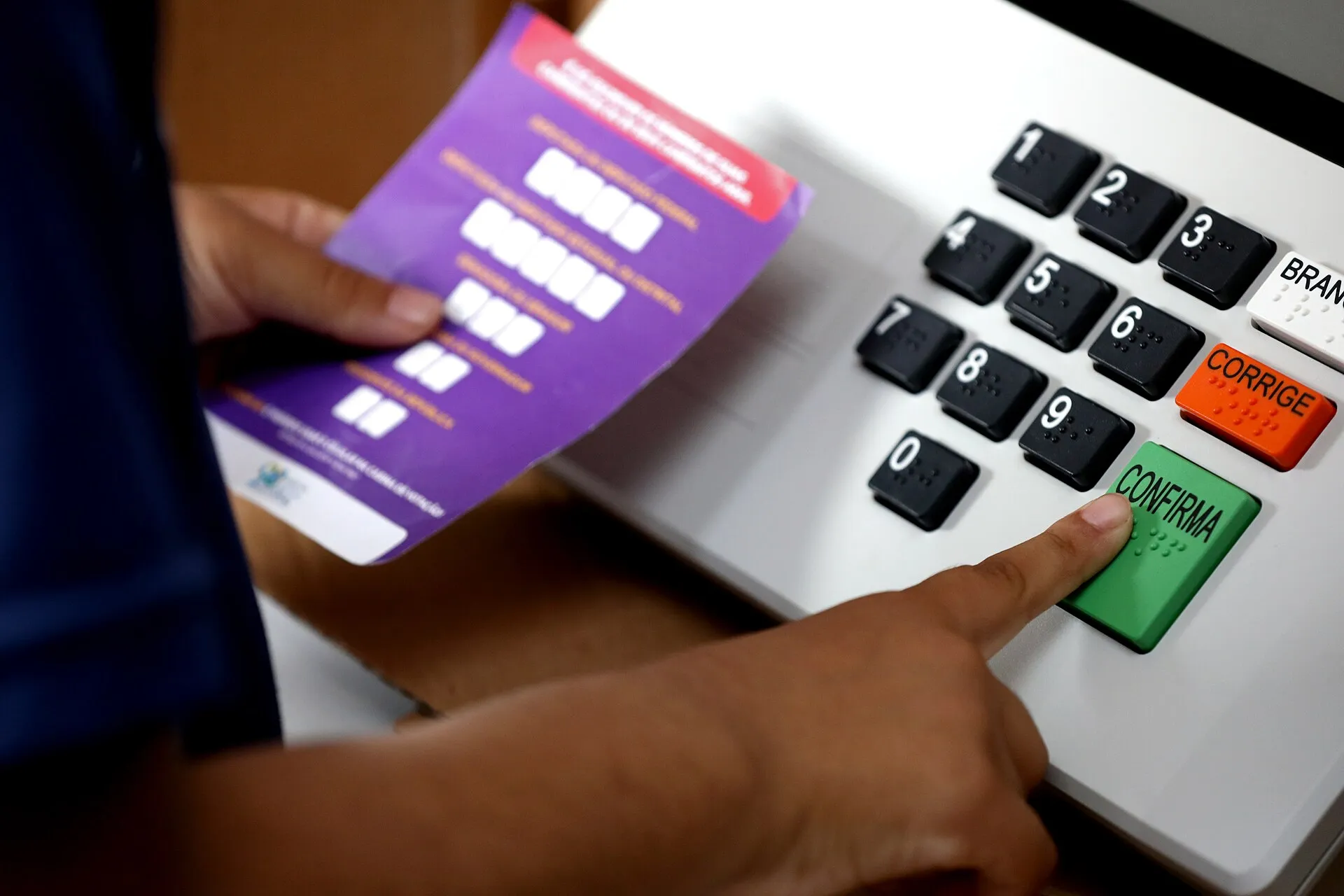 Mão de uma pessoa aponta a tecla Confirma de uma urna eletrônica, ao lado de um cartão de demonstração roxo com campos de voto