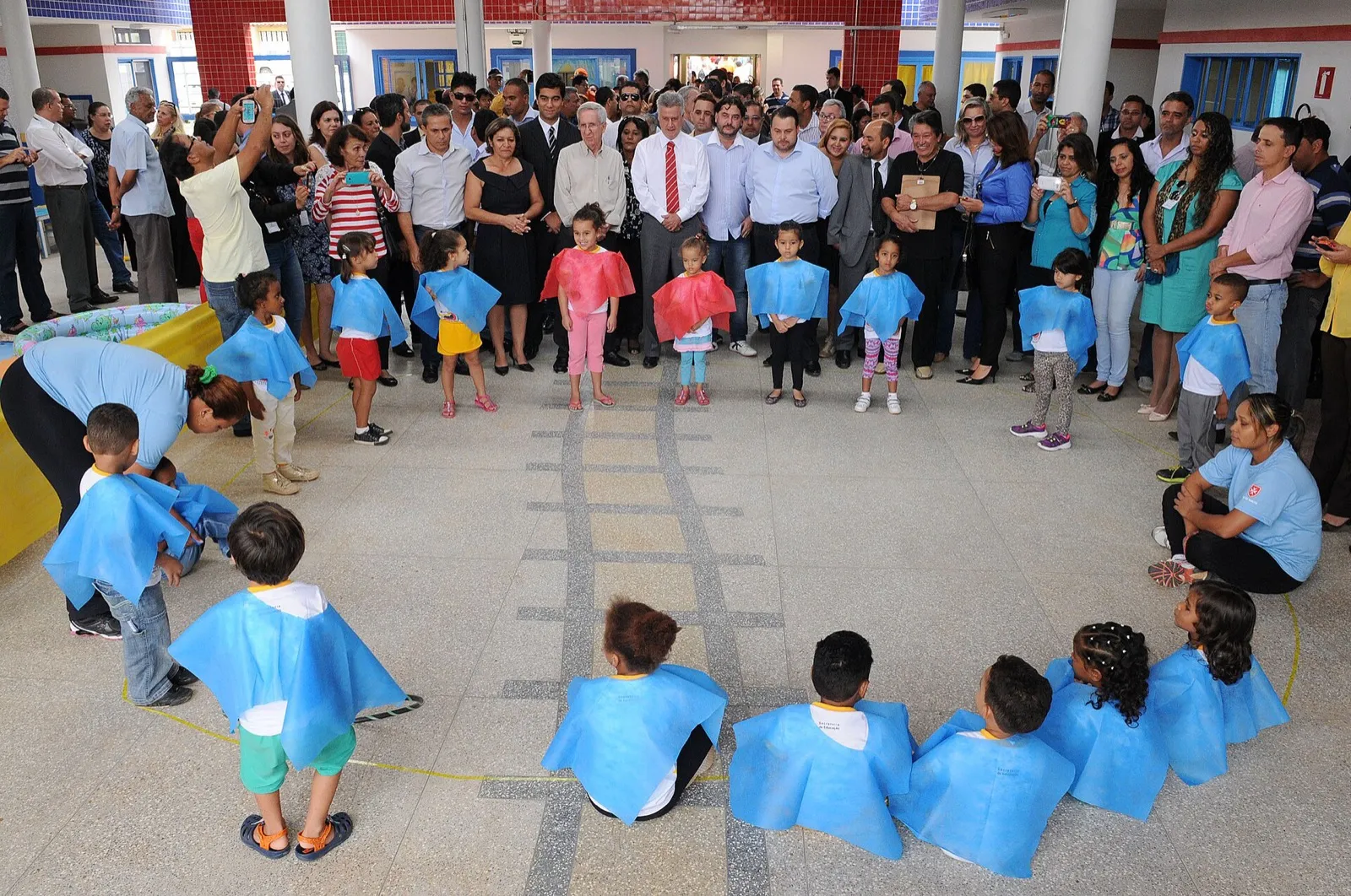 Crianças pequenas de capas azuis formam uma roda em uma creche pública de Sobradinho, no Distrito Federal, com adultos observando ao redor