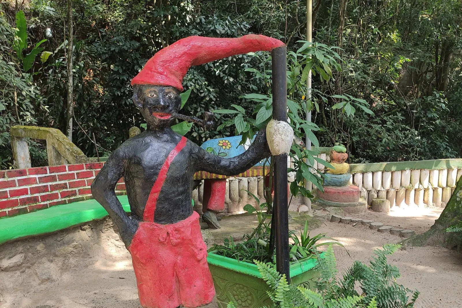 Escultura colorida de Saci Pererê, com boina vermelha, cachimbo na boca e um poste preto na mão, em um jardim com samambaias