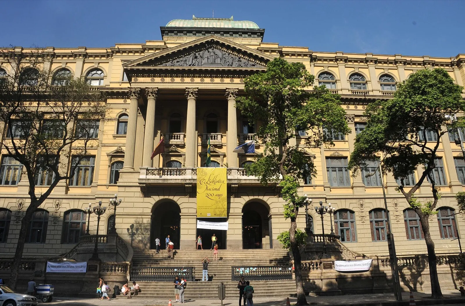 Fachada amarela e neoclássica da Biblioteca Nacional, no Rio de Janeiro, com faixa de 200 anos pendurada sobre a entrada e pessoas na escadaria