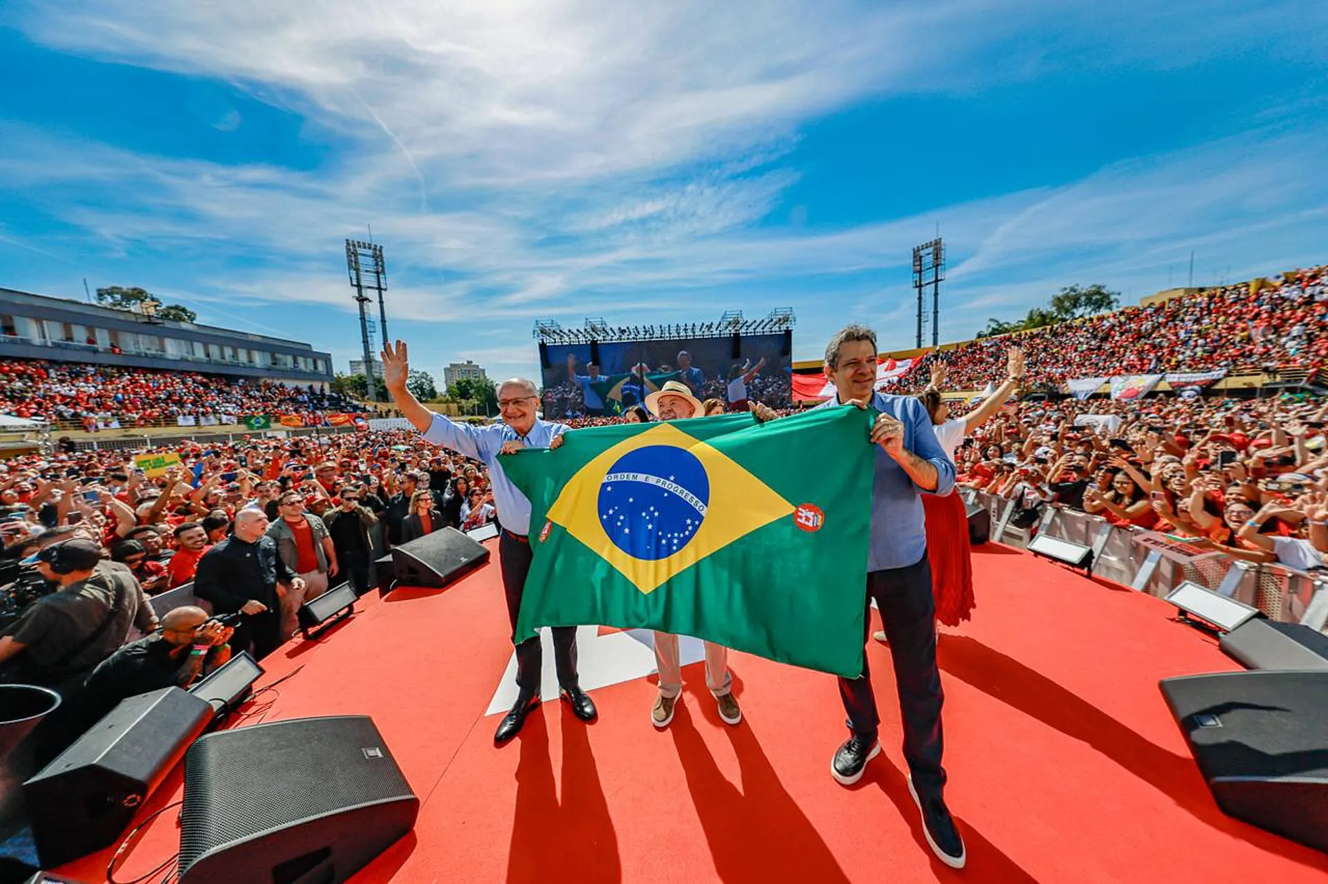 Três homens seguram uma bandeira do Brasil no palco de um estádio lotado, durante o lançamento da campanha de Lula