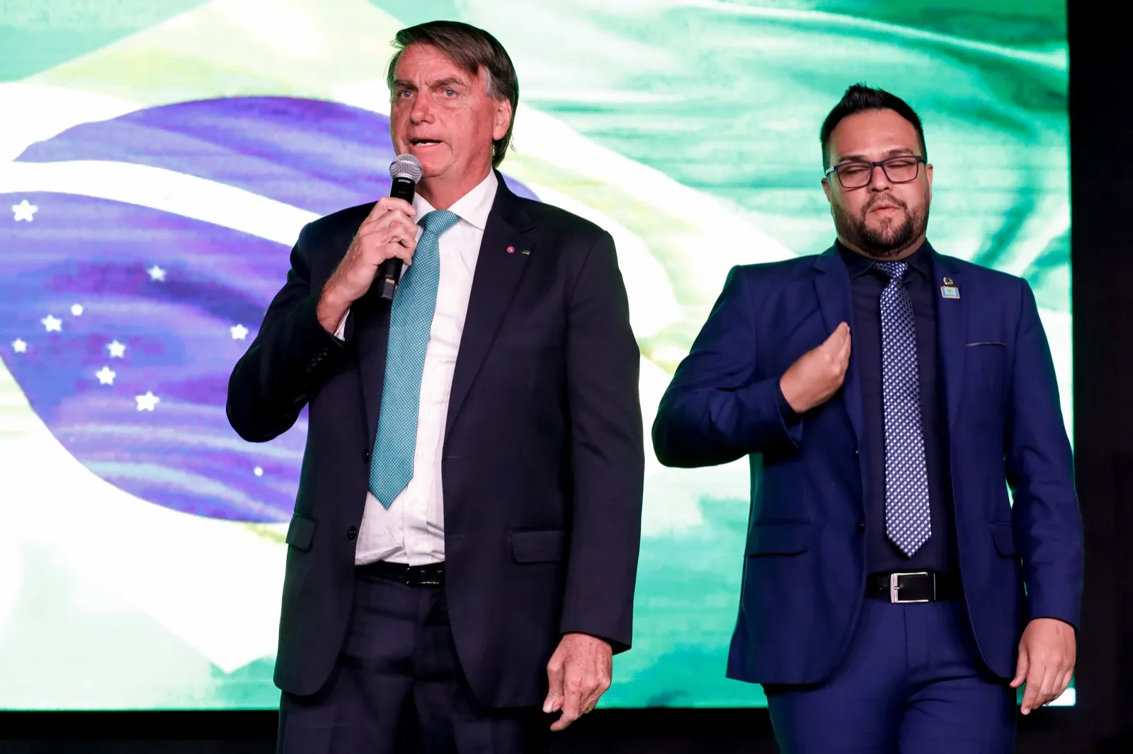 Jair Bolsonaro fala ao microfone em um palco, ao lado de um intérprete de Libras, diante de um telão com a bandeira do Brasil