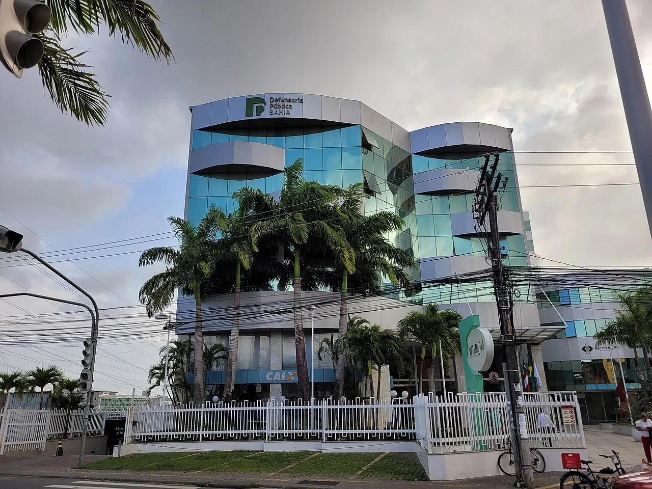 Prédio de vidro azul e painéis prateados com o logotipo da Defensoria Pública da Bahia, atrás de palmeiras, em Salvador