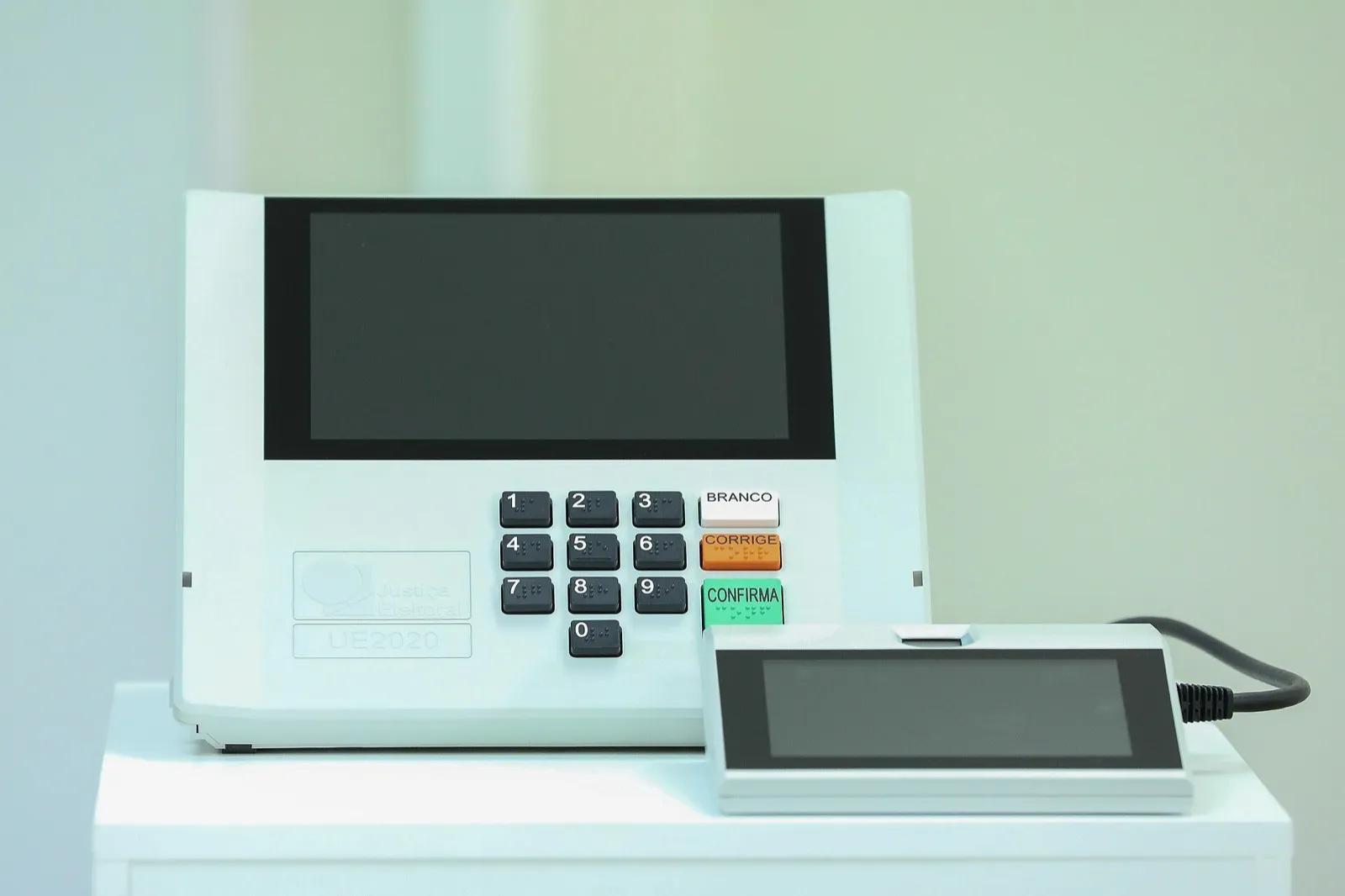 Urna eletrônica branca com tela, teclado numérico e as teclas Branco, Corrige e Confirma, ao lado do terminal do mesário