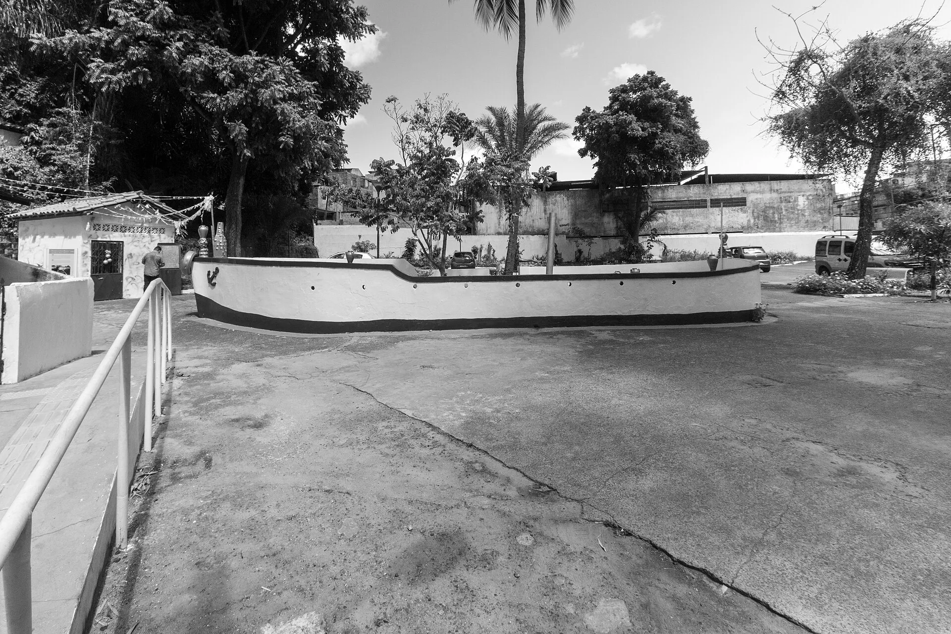 Tanque branco em forma de barco no pátio do Terreiro da Casa Branca, em Salvador, com árvores, um muro e um pequeno prédio ao fundo.